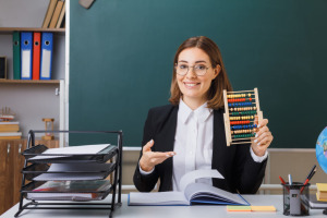 young-woman-teacher-wearing-glasses-sitting-school-desk-front-blackboard-classroom-using-abacus-explaining-lesson-happy-positive-smiling-1024x683